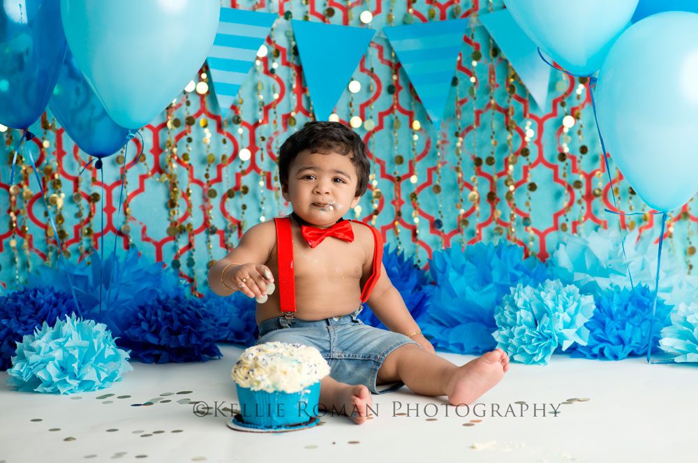 For The Love of Cake Smash one year old boy with frosting on face and fingers. Wearing red bow tie and suspenders. Infant of red and blue backdrop with gold beads, blue balloons, tissue paper pom poms and blue banner