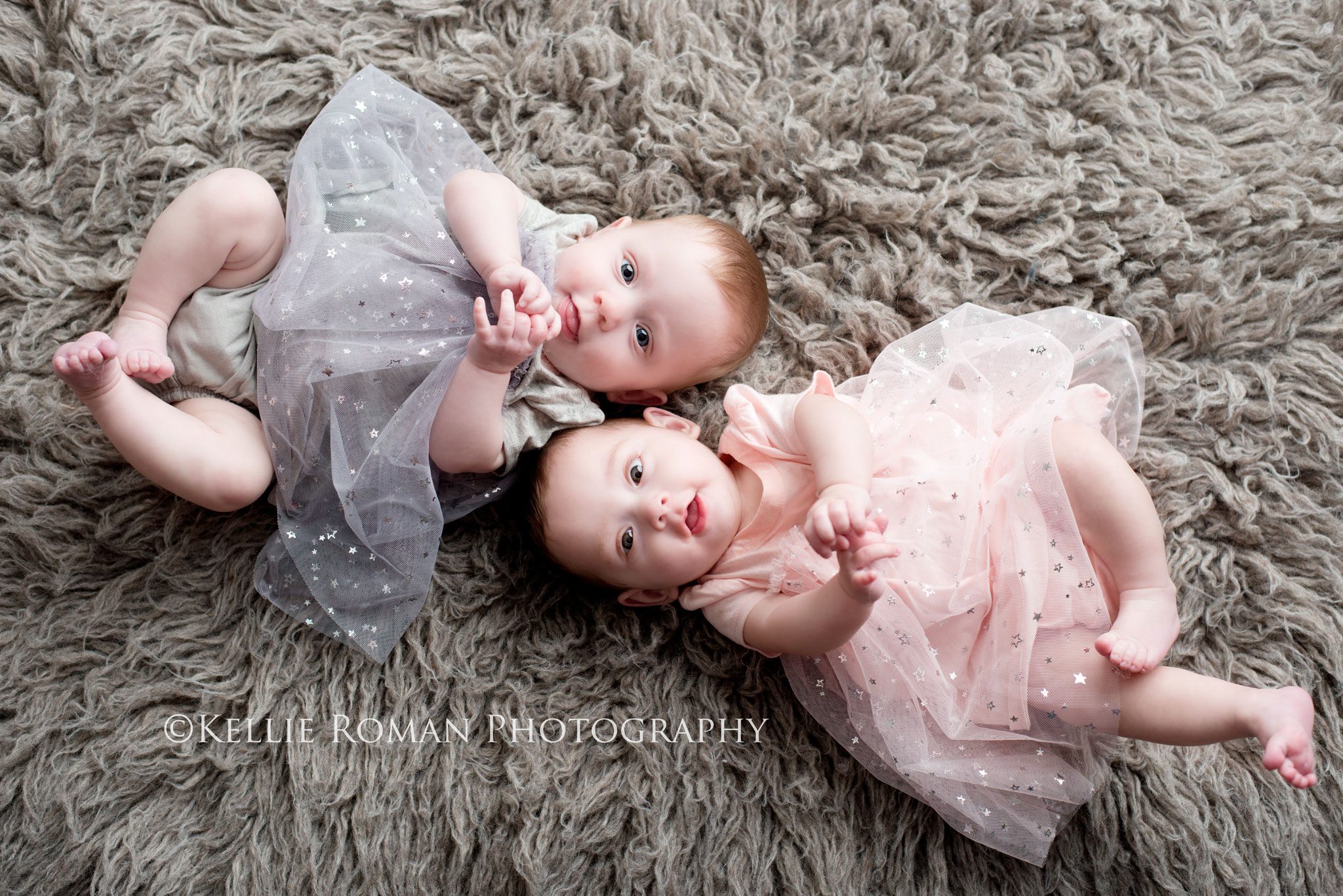 Milwaukee Twin Photographer six month old twin girls laying side by side onto of grey rug both hands and feet in the air
