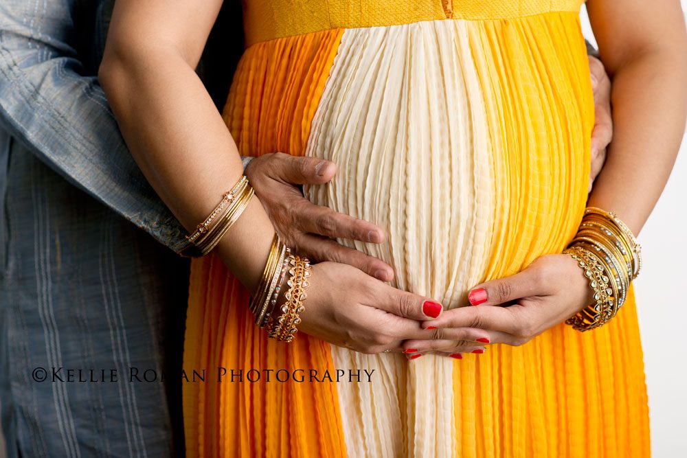 For The Love of Cake Smash close up of husband and wife putting hands on pregnant wife's belly. Couple wearing orange and grey with gold jewelry.