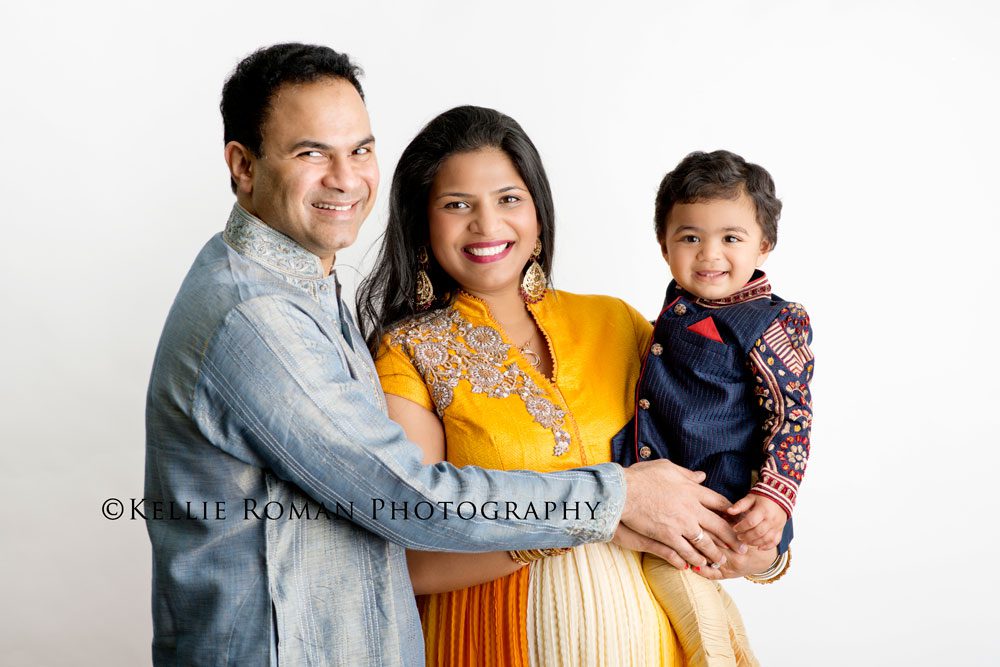 For The Love of Cake Smash family photo infant of white backdrop. Family wearing traditional Indian outfits, mother holding one year old son.