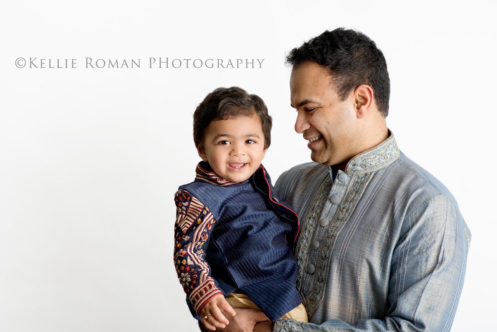 For The Love of Cake Smash father and toddler son infant of white backdrop. Son looking at camera and father looking at son. Both are smiling and wearing blue and grey clothing.