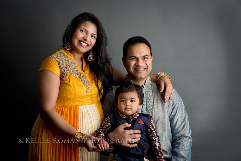 For The Love of Cake Smash family of three infant of dark grey backdrop. Father sitting on stool holding toddler son, mother standing while hold son's hand. Wearing traditional Indian clothing.