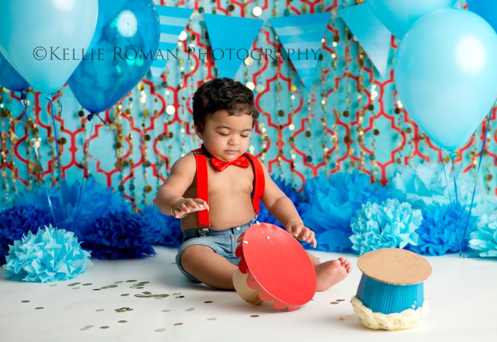 For The Love of Cake Smash toddler boy wearing red bow tie and suspenders infant of red and blue backdrop with gold beads. Blue balloons, tissue paper pom poms. Cake tipped over on floor.