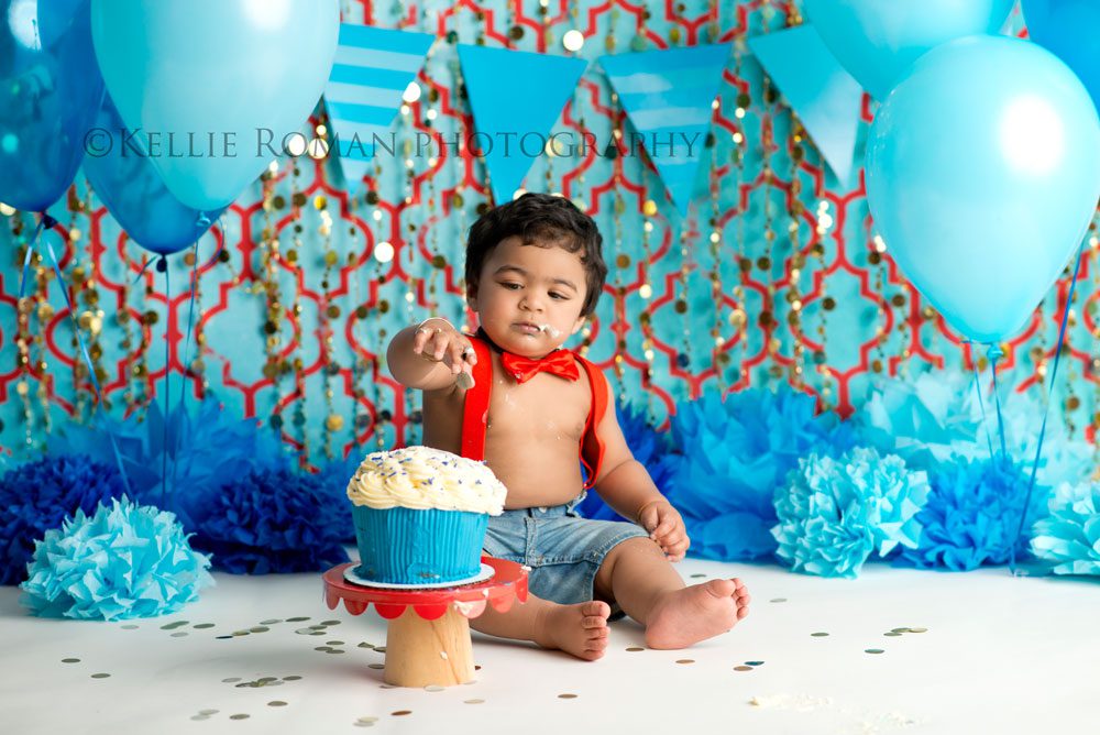 For The Love of Cake Smash one year old boy looking at blue and white cake onto of red cake stand. Infant of blue red and gold backdrop balloons and pom poms