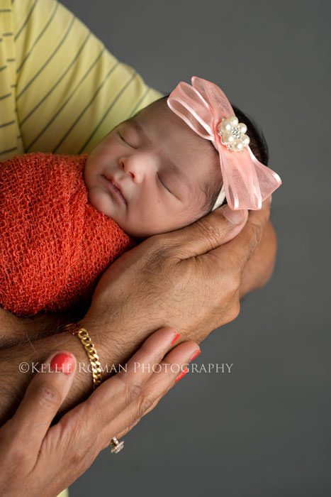 flower bonnet close up of grandparents hands on newborn grand daughter sleeping with pink headband and orange swaddle wrap