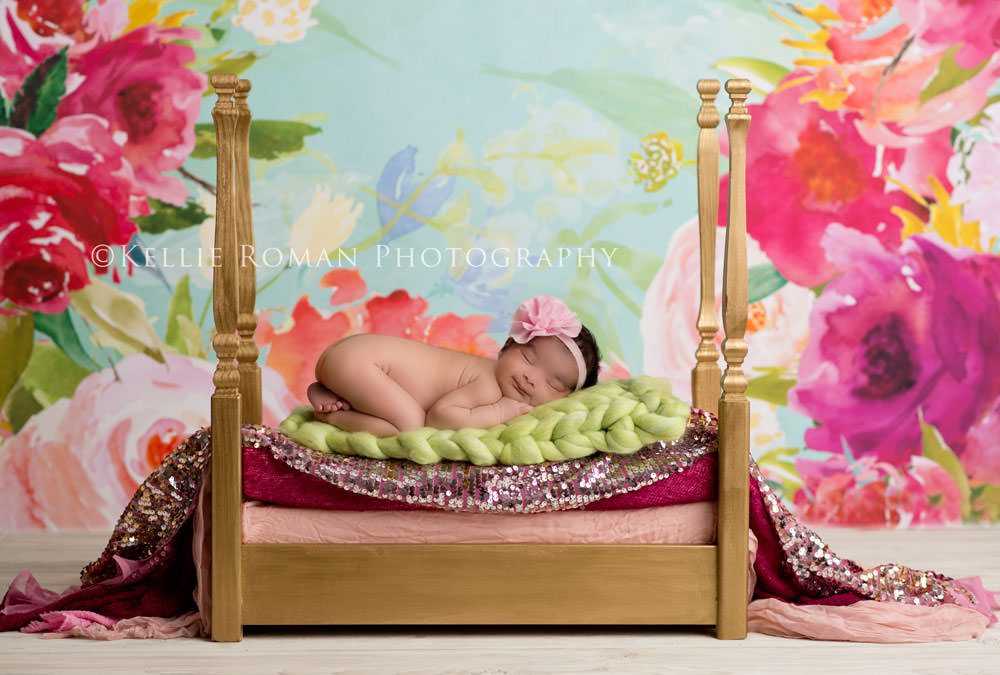 Newborn Bonnet. Newborn girl laying on top of gold bed with fabric layers. Floral backdrop with pink red green blue and gold colors. Newborn girl smiling.