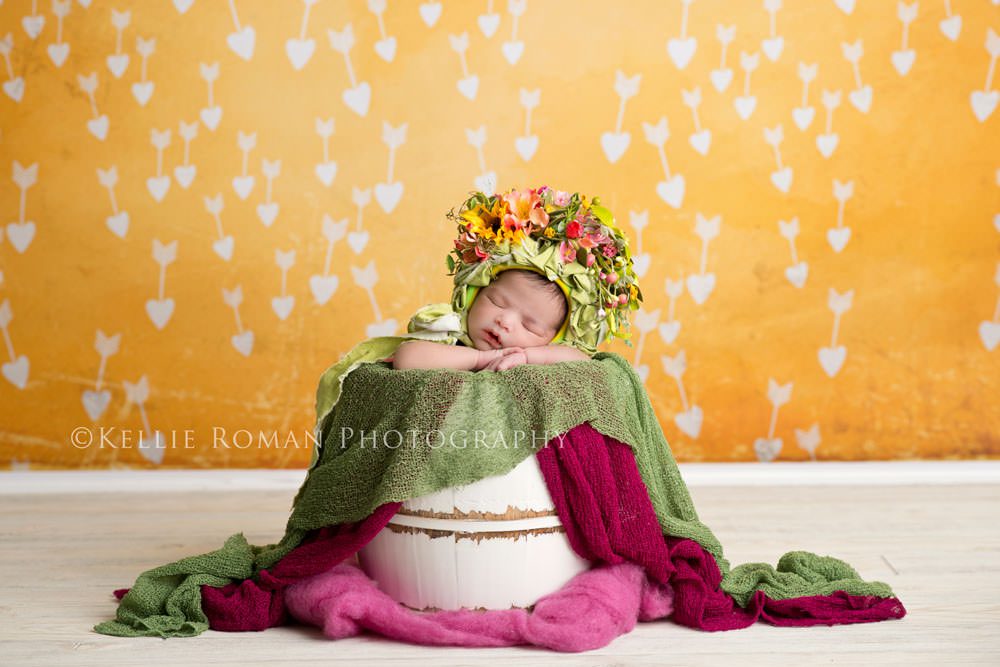 Flower Bonnet. Infant posed in white bucket. Chin on hands sleeping with bonnet made of color flowers. White wood floor with yellow backdrop. Pops of dark pink colors.