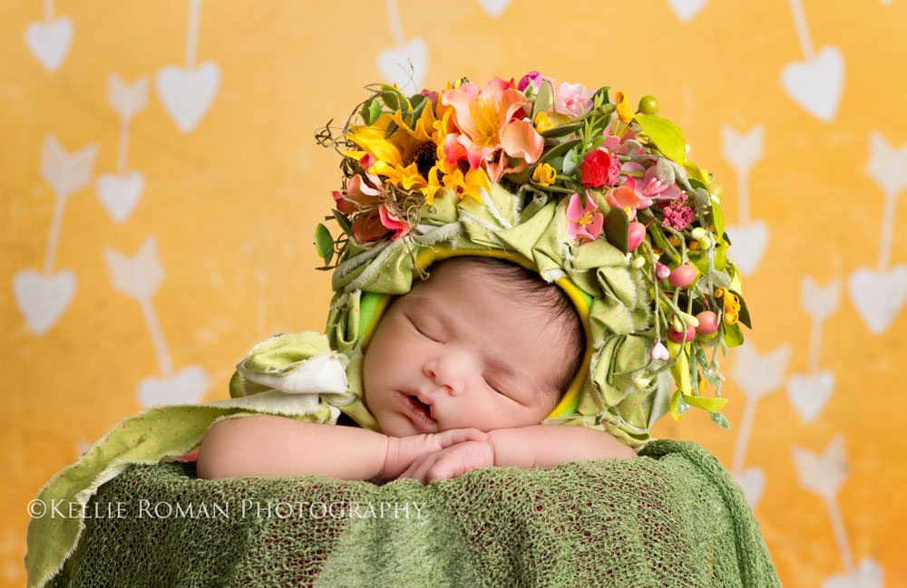 Flower Bonnet. Newborn girl posed in bucket with chin on hands. Wearing bonnet with pink yellow green orange flowers. Yellow backdrop with hearts.