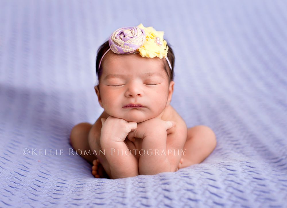 Flower Bonnet. Newborn girl onto of purple fabric on bean bag poser. Froggy pose. Chin on hands and toes visible. Wearing yellow and purple headband