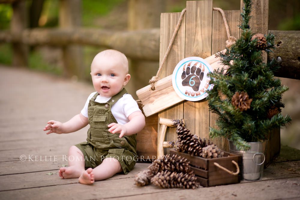 outdoor little man baby boy sitting on wood bridge in overalls with pinecones trees and a sign