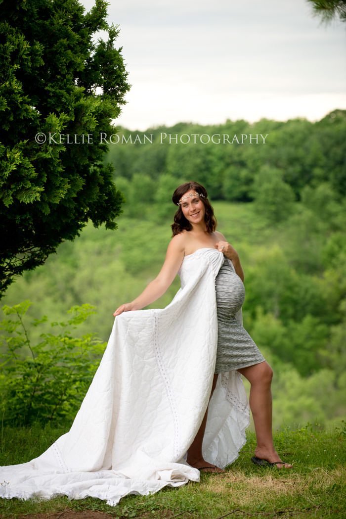 vintage style photo shoot maternity model wrapped in blanket with headband standing in front of green trees 