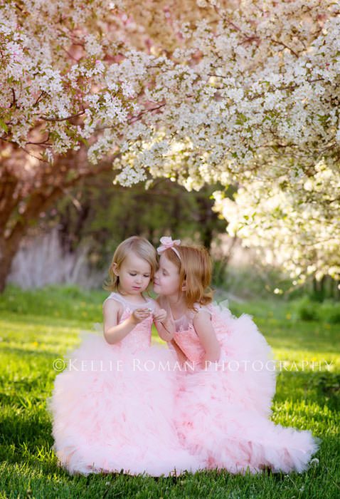 in full bloom sisters standing under flowering trees wearing ballgowns one is crouched down to kiss the other girl