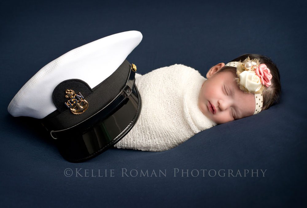 little sailor girl newborn girl wrapped in white swaddle on top of navy fabric with fathers naval officer hat 