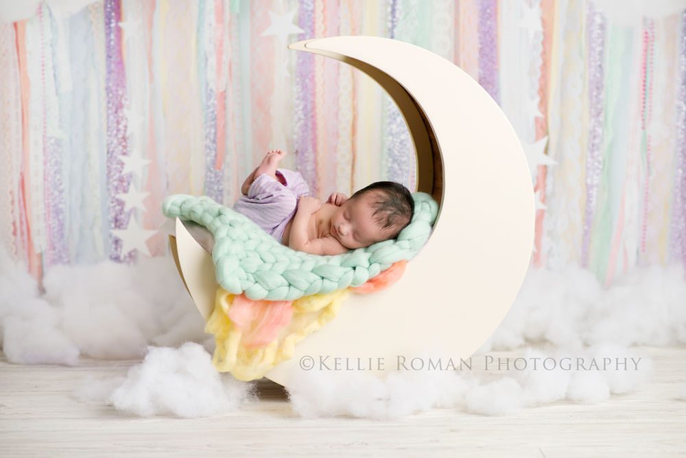 little sailor girl newborn girl wearing purple pants posed on back in moon prop with clouds around her and a pastel ribbon backdrop