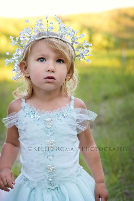 the great lakes toddler girl wearing silver jewel crown and blue beaded dress looking up standing in park