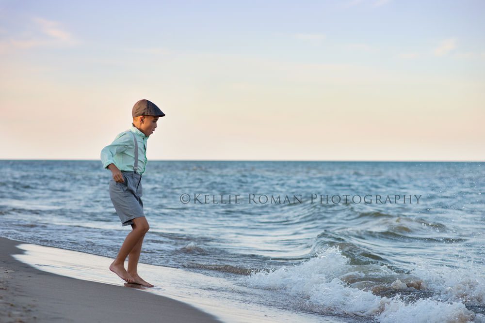 the great lakes boy hopping excitedly on sand while wave rolls in wearing grey shorts suspenders and hat