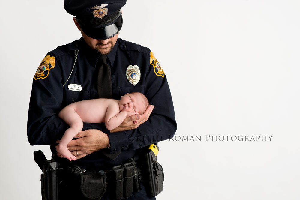 three big sisters naked newborn boy laying on belly in dads arms dad is wearing police uniform infant of white backdrop
