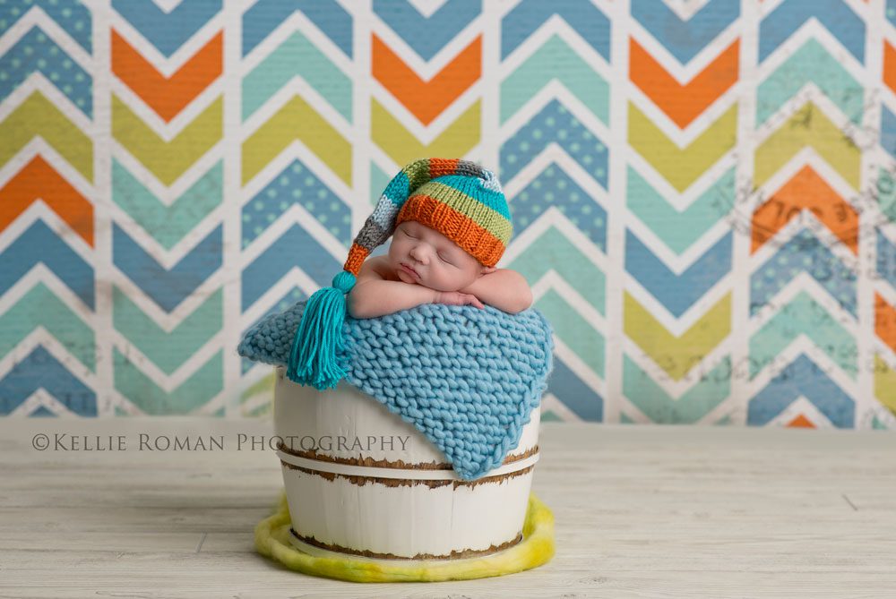 three big sisters baby posed in bucket with orange green teal hat on and infant of a chevron backdrop