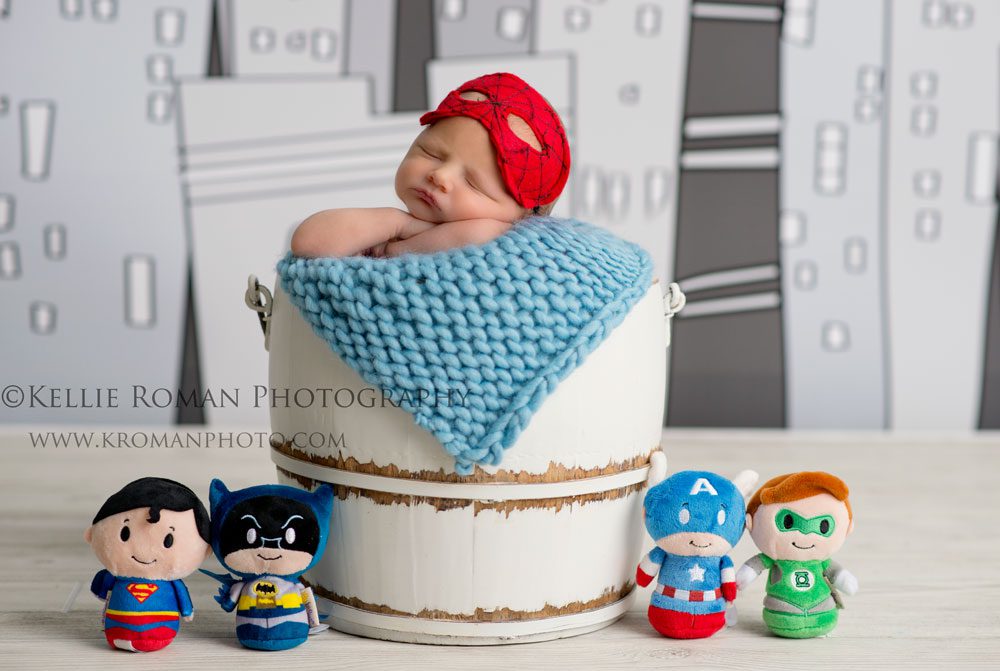super hero newborn posed in white bucket with super hero stuffed animals around him he has a spider man mask on