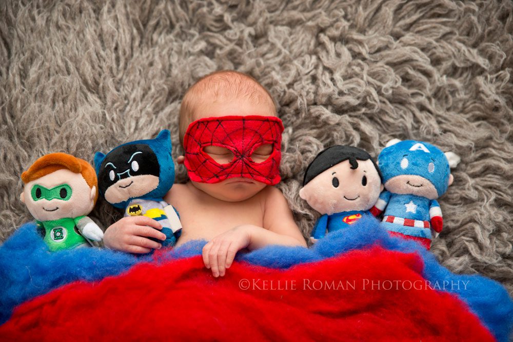super hero newborn boy laying on grey fur rug with spider man mask on and holding small super hero stuffed animals