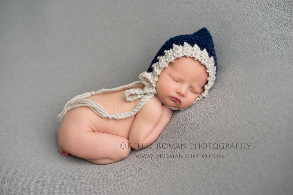 super hero sleeping infant boy on his stomach with navy and grey bonnet on onto of grey blanket