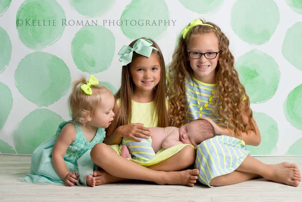 three big sisters three girls in front of polka dot backdrop baby brother is on girls lap one sister look at baby
