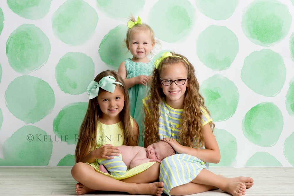 three big sisters three girls posed in front of green polka dot backdrop with newborn brother on lap