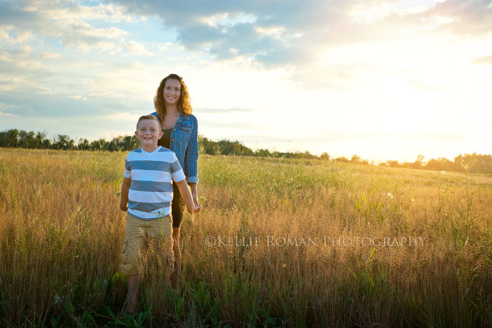 mother and son session mother holding sons hands while he's in front of her they are standing in a field of tall grass in the sunset with blue cloudy sky