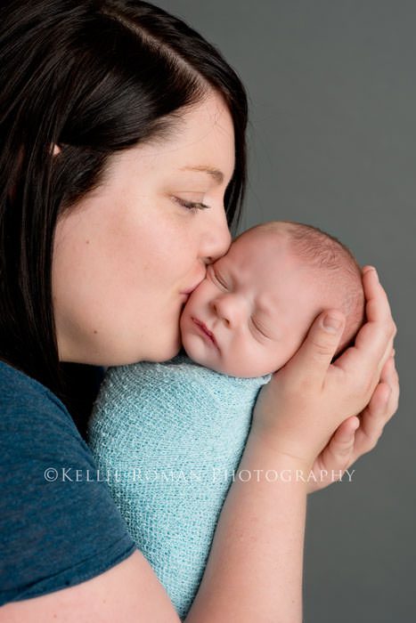 newborn photography mom holding newborn boy swaddled in blue wrap and kissing his cheek dark grey backdrop