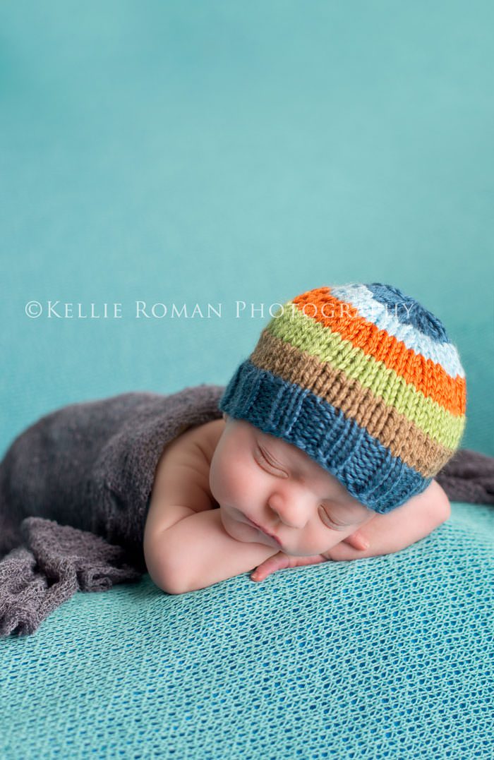 newborn photography baby boy sleeping with chin posed on top of arms he he laying on his belly onto of a blue blanket with a striped hat on and a grey wrap over his diaper