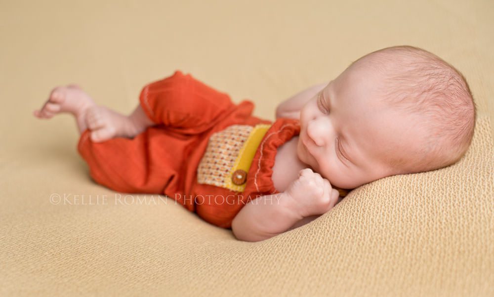 newborn photography baby boy posed on back sleeping onto of yellow blanket wearing orange and yellow romper