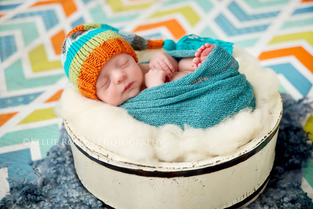 newborn photography baby boy wrapped up in swaddle with toes and hands sticking out sleeping on his back in a white bucket wearing striped hat that matches the chevron back ground 