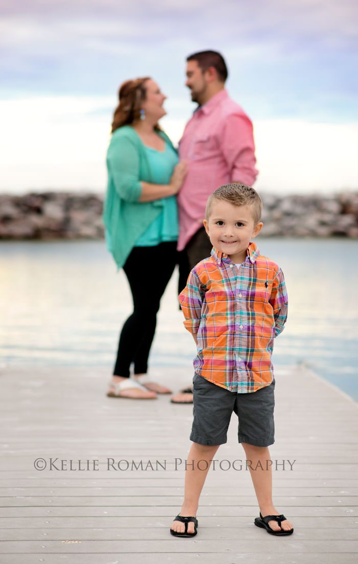 family photography couple in background looking at each other while son is in the foreground smiling taken on a pier outside by water