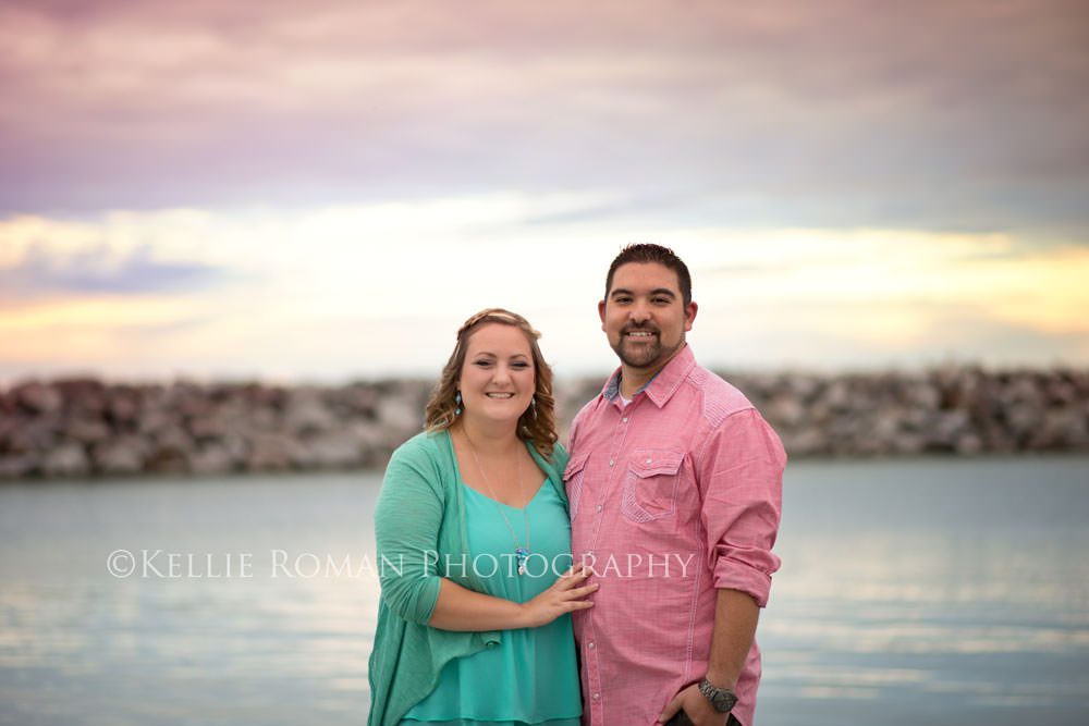 family photography husband and wife standing infant of water on a pier looking at camera sky is pastel colors