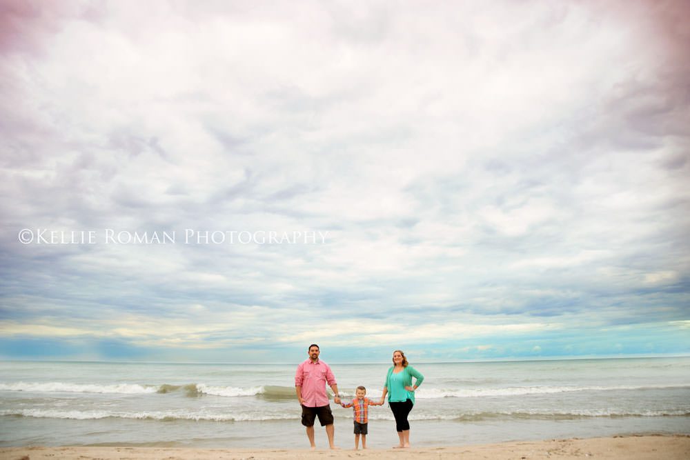family photography husband and wife standing on beach hold sons hand with pastel clouds in the sky
