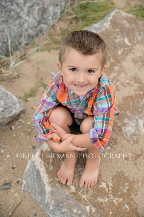 family photography boy sitting on rock on a beach with orange plaid shirt on smiling at camera