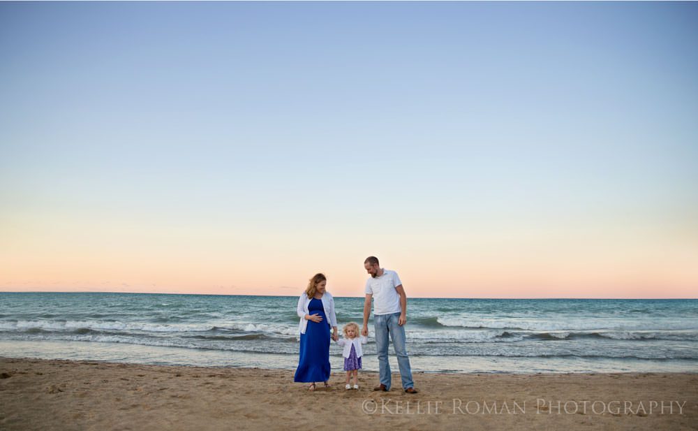 family photos husband and wife with toddler daughter all holding hands standing on beach with pastel colored sky during dusk