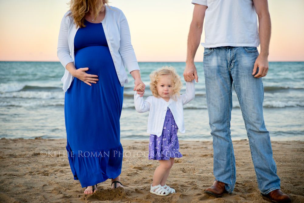 family photos husband wife and toddler daughter standing on a beach holding hands close up of toddler with mother holding pregnant belly