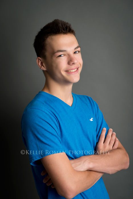 senior pics boy standing in studio in front of grey backdrop with blue shirt on