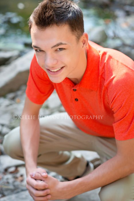 senior pics boy squatting onto of rocks looking up and camera and smiling he has an orange shirt on