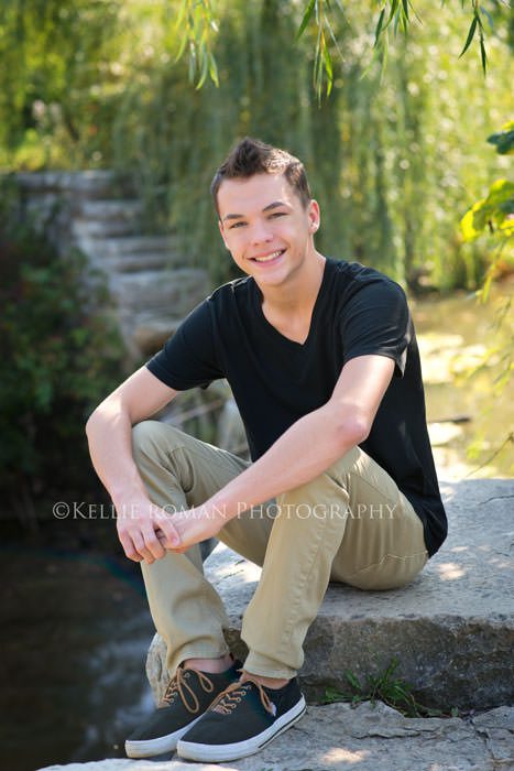senior pics boy with black shirt on and khakis sitting on rocks in a park 