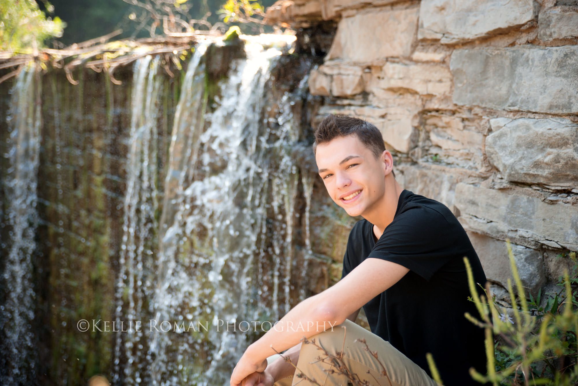 boy sitting on rocks in front of waterfall with black shirt on smiling at camera during senior pics