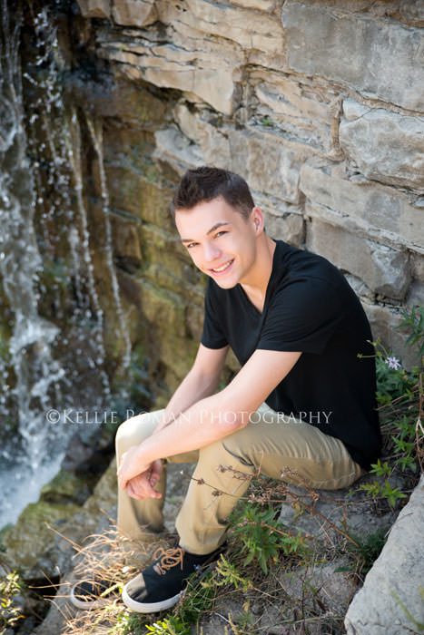 boy sitting on a rock in front of water in a black shirt during senior pics