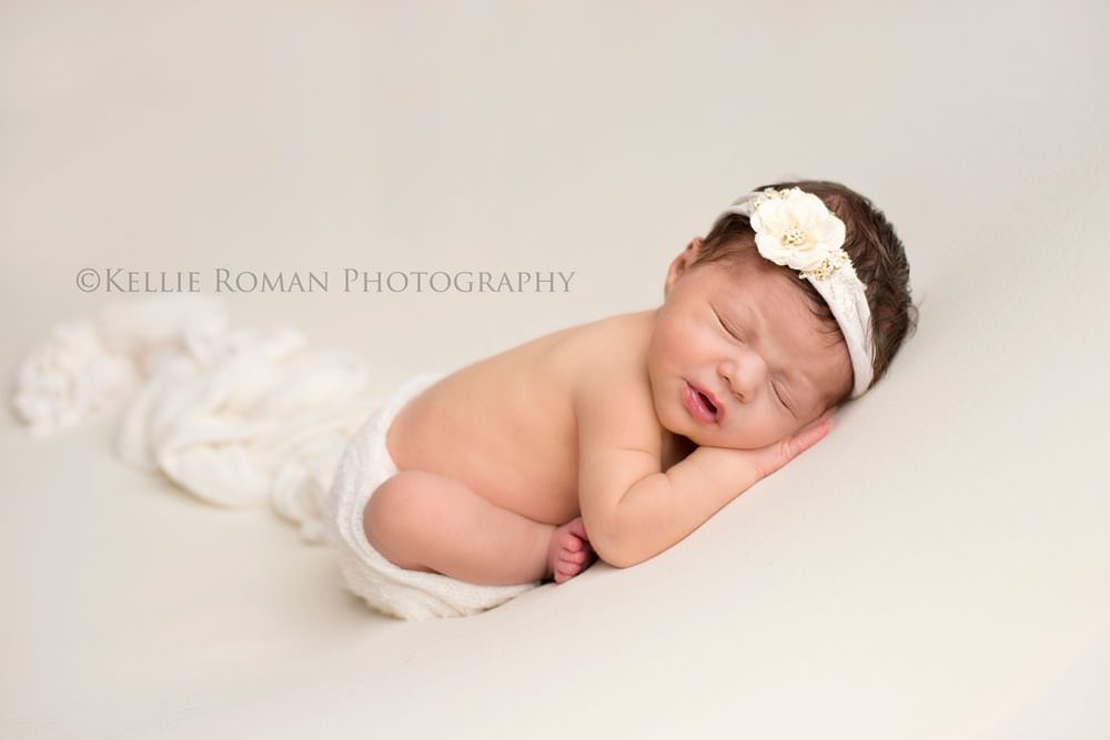 newborn session with baby girl in taco pose onto of cream colored blanket. She has a white wrap around her butt and a white flower headband on. 