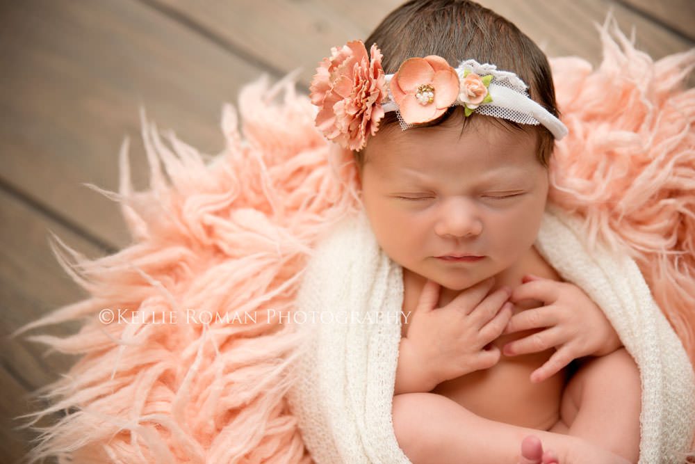 newborn session with baby girl in white wrap onto of a pink fur in a bucket. She has a pink flower headband on and has her hands resting on her chest.
