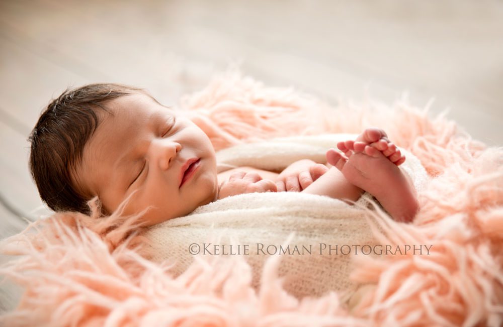 Newborn session. Infant girl in white wrap in a bucket onto of pink fur. Lighting is coming from behind her. She has dark brown hair.