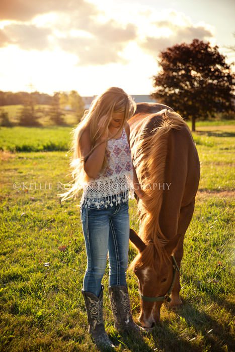 high school senior photos young girl standing in field with horse with sun shining through their hair and clouds in the sky