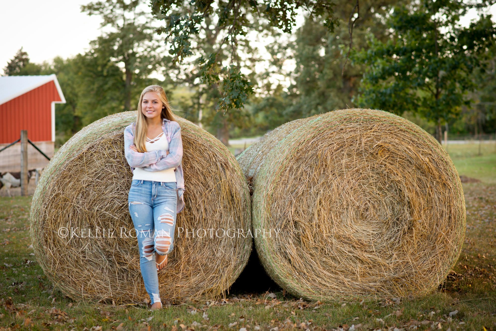 high school senior photos senior girl standing outside on family farm. She's leaning against giant hay bails. She has long blonde hair, ripped jeans on and a flannel shirt