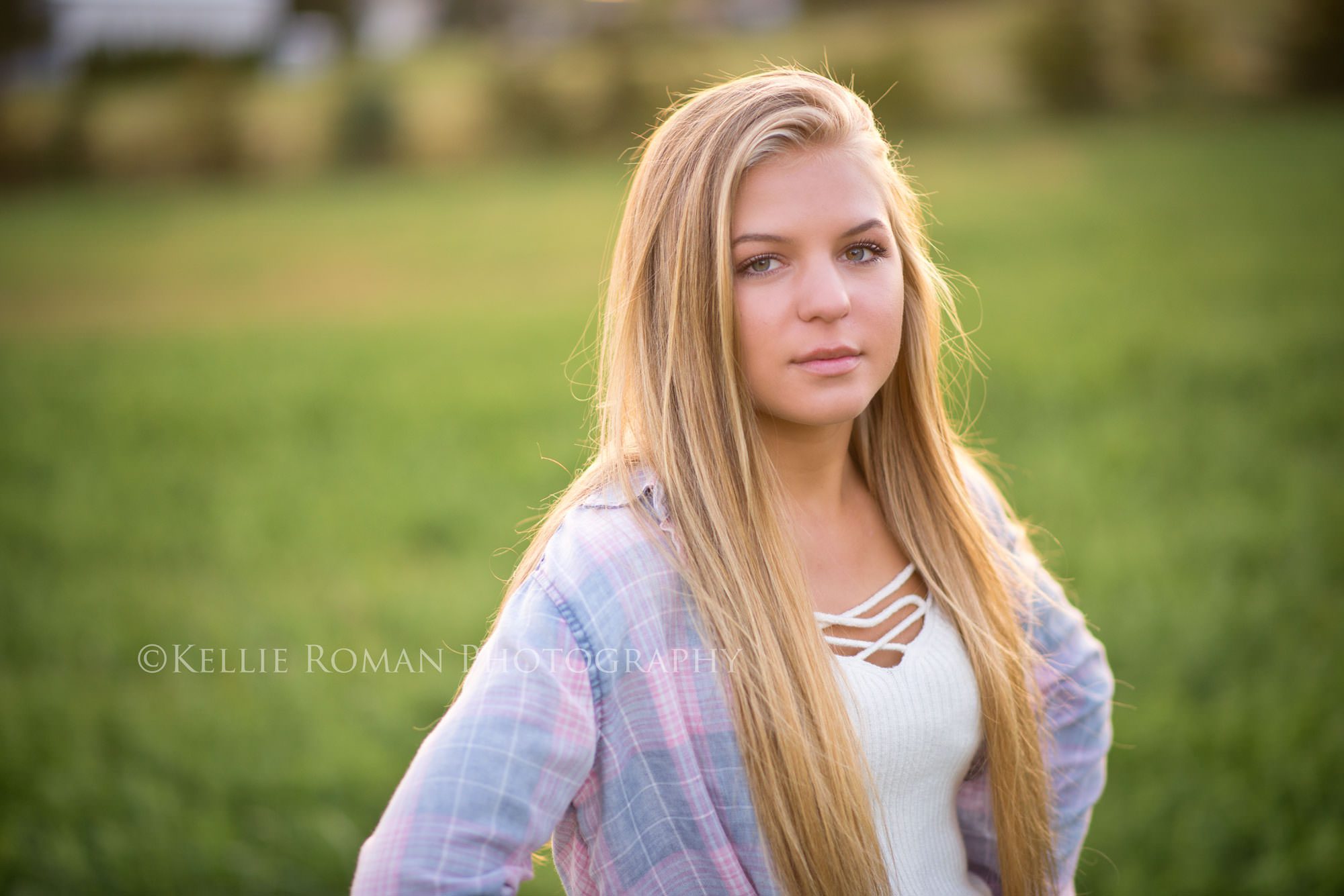 high school senior photos senior girl with serious face standing in field of grass outside on family farm. She has long blonde hair and a flannel shirt on