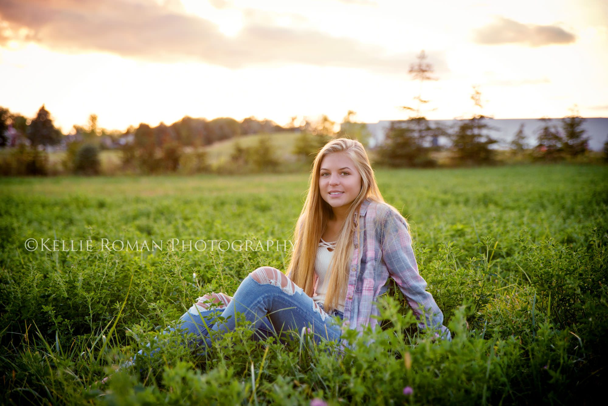 high school senior photos senior girl sitting in green grass within a big field clouds in the sky and she is wearing a flannel shirt and jeans